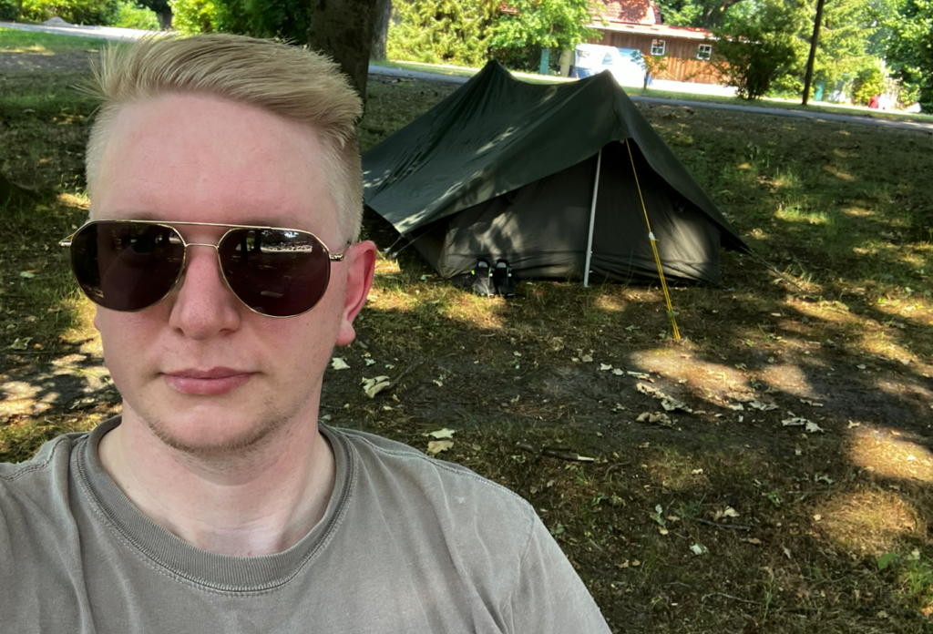 Selfie von Tjark Siegfriedson mit dem aufgebauten Zelt im Hintergrund. Ein Tente F1 von der französischen Armee